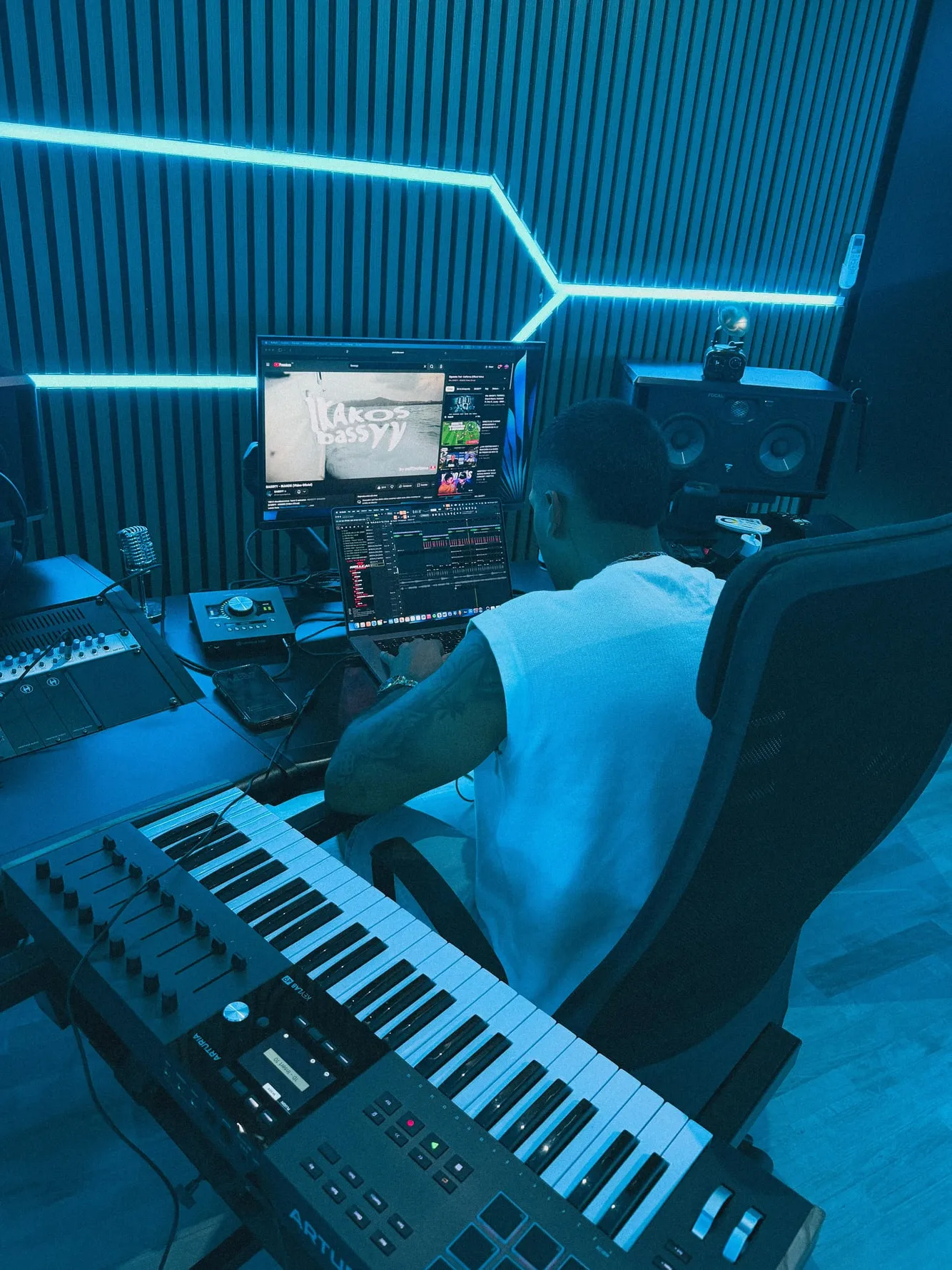 VaronW de espaldas en su estudio, produciendo frente al teclado y los monitores, con luces de neón azul